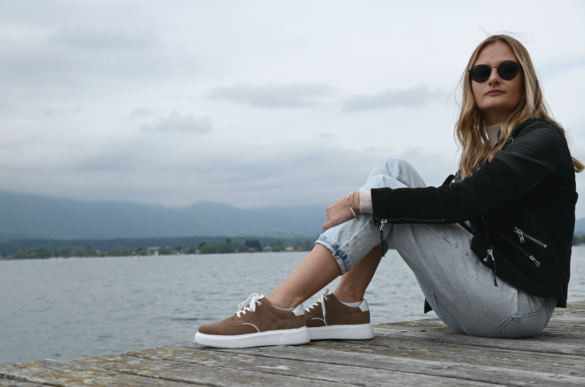 Frau mit braunen CYBRUS Sneaker sitzt auf einem Holzsteg. 